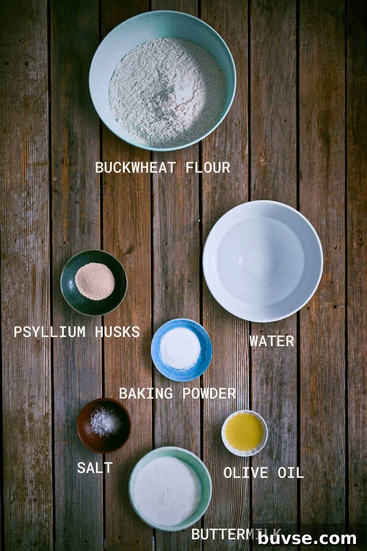 Gluten-Free Buckwheat Pita Bread Recipe 4 Seven bowls of ingredients for baking-buckwheat flour, psyllium husks, baking powder, salt, buttermilk, olive oil, and water-are arranged on a wooden surface, each labelled with its name.