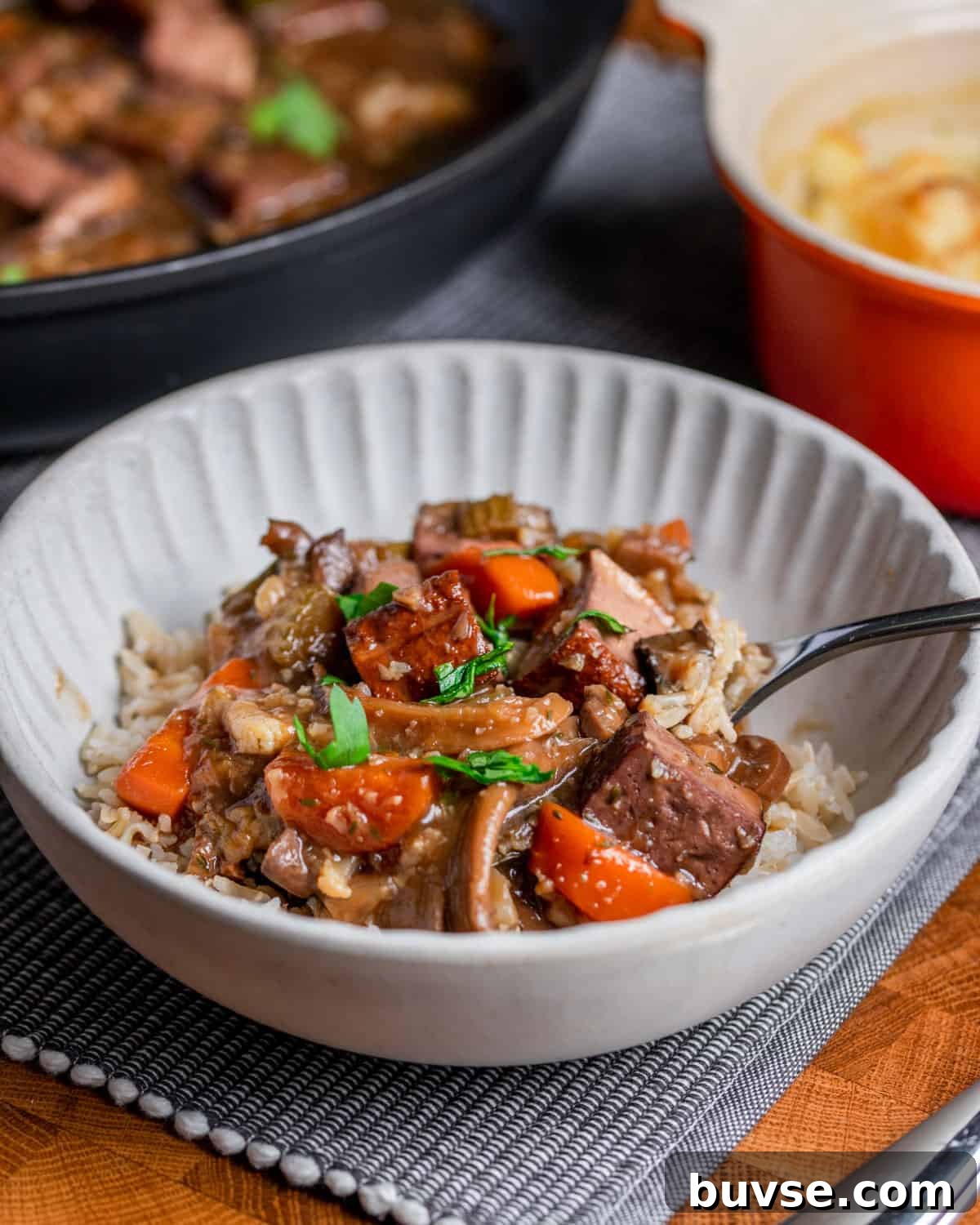 Photo of a bowl of vegan stew over brown rice, set on a table mat with vegan cauliflower cheese in the background and the pan of stew.