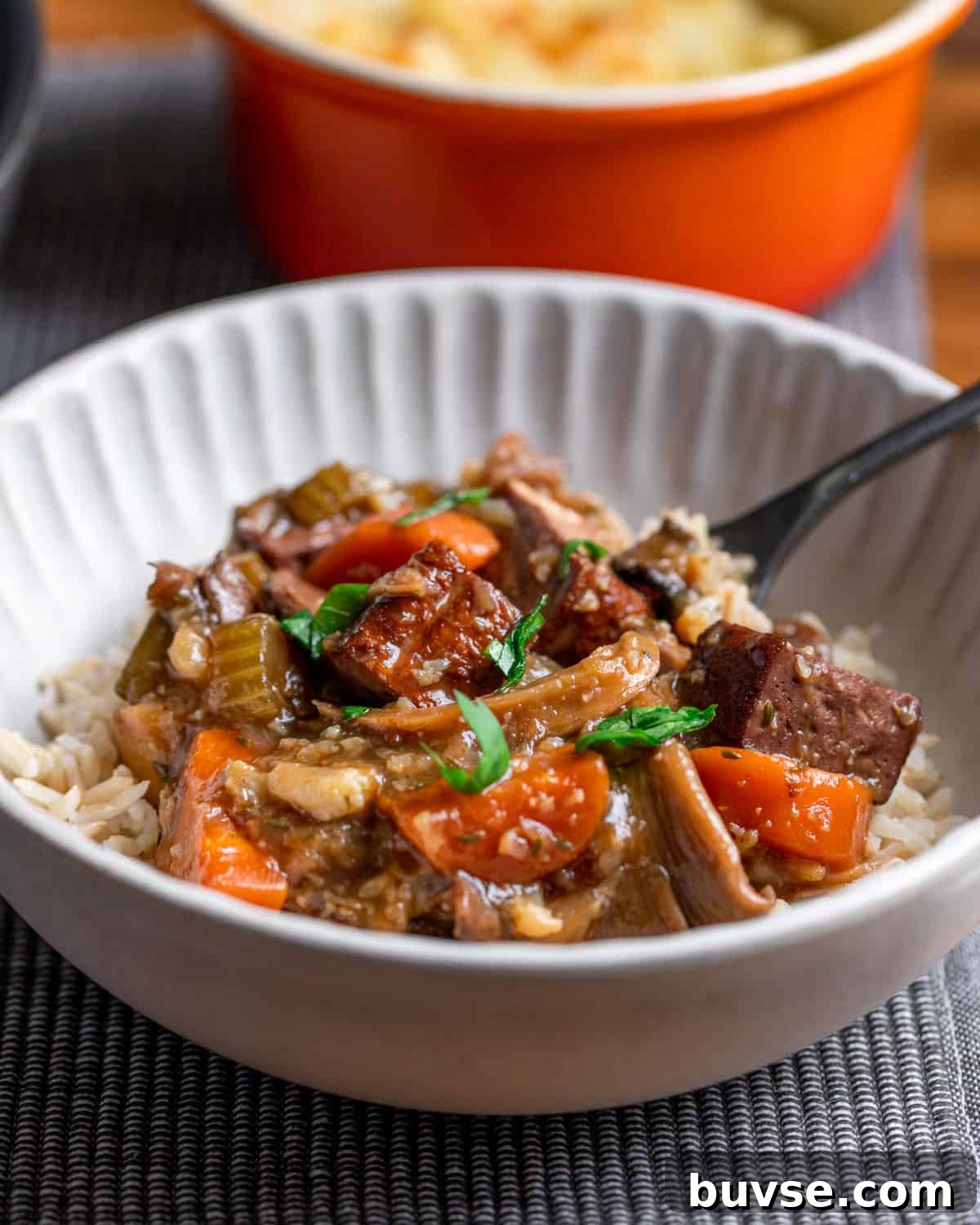 Photo of a bowl of vegan stew served with brown rice and a spoon, set on a grey mat with an orange pot in the background.