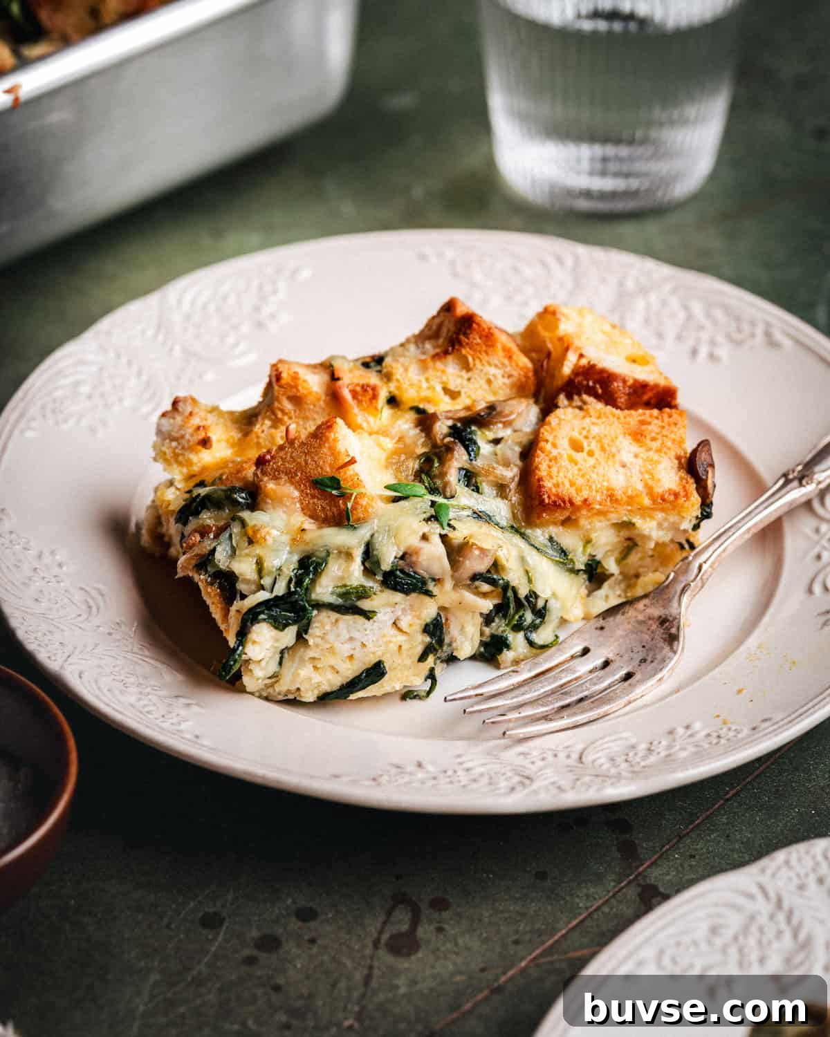 A slice of savory bread pudding with spinach, cheese, and mushrooms sits on a white plate with a vintage fork. A glass of water and more casserole are in the background.