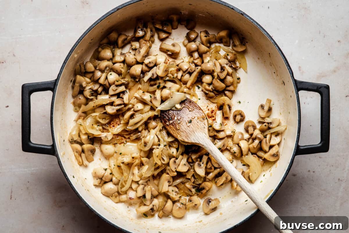 A large pot filled with sautéed mushrooms and onions, being stirred with a wooden spoon on a light countertop.