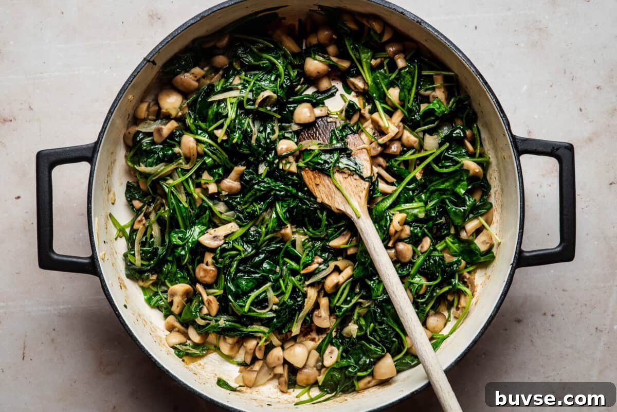 A large pan filled with sautéed spinach and chopped mushrooms, mixed together and stirred with a wooden spoon on a light-colored surface.