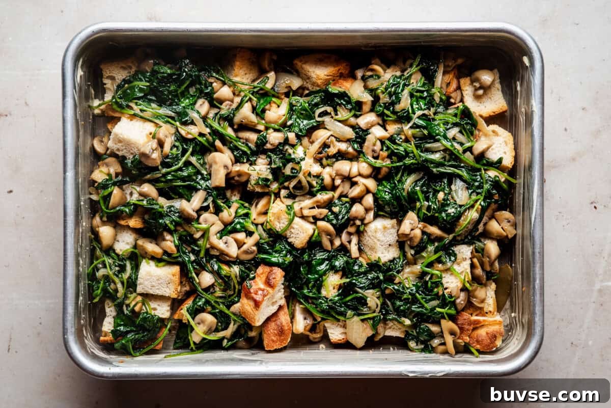 A baking dish filled with sautéed spinach, mushrooms, onions, and cubes of toasted bread, arranged evenly and ready to be baked.