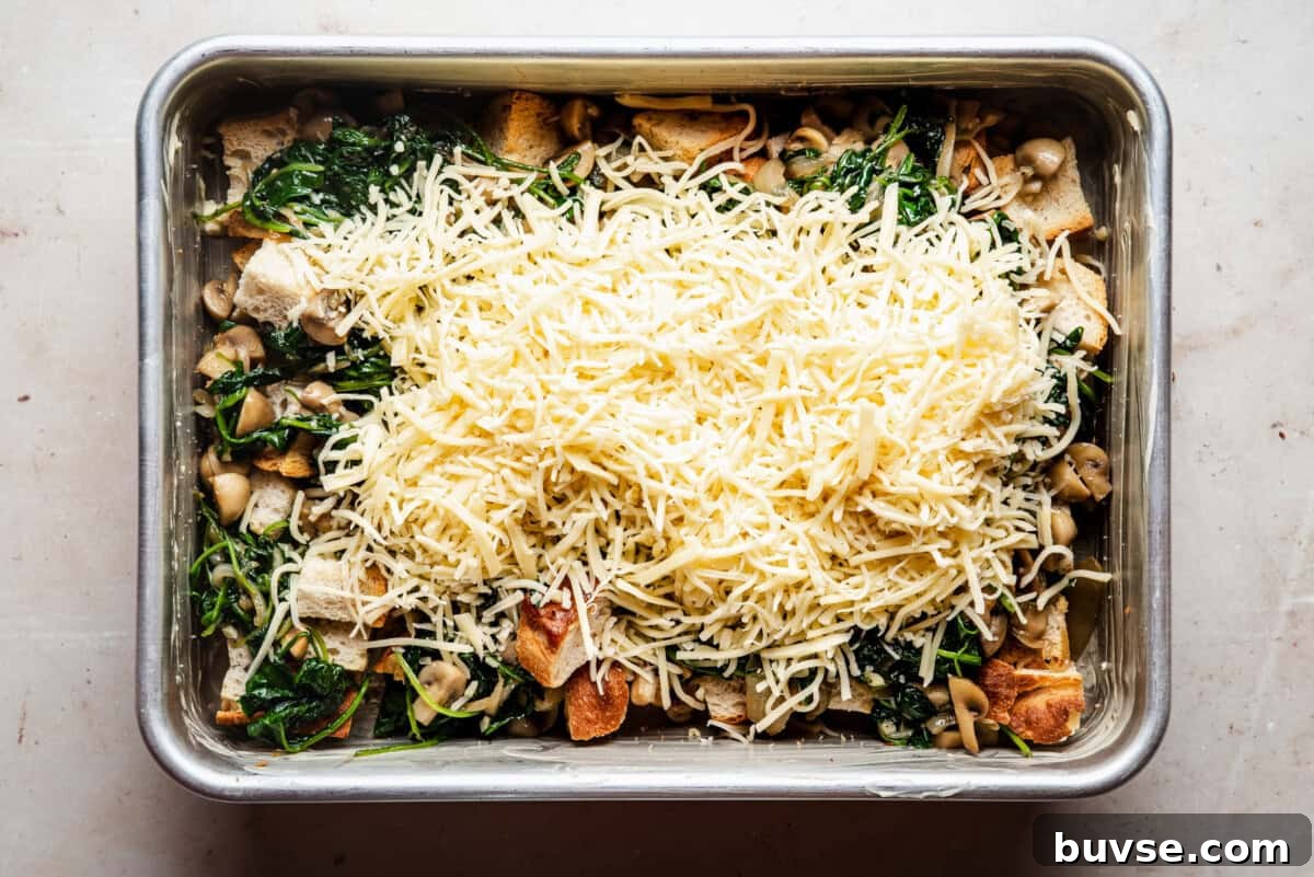 A rectangular metal baking dish filled with a mixture of sautéed spinach, mushrooms, bread cubes, and topped with a generous layer of shredded cheese, ready to be baked.