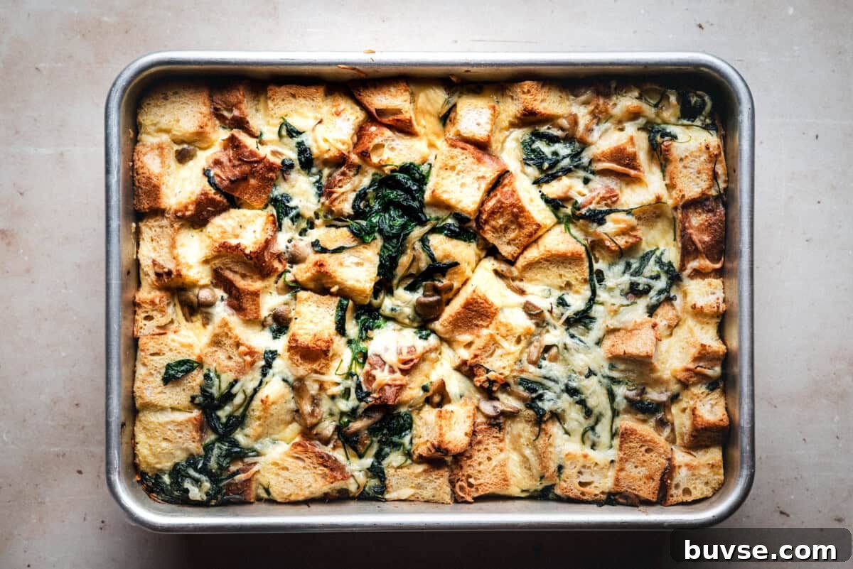 A baked bread pudding in a rectangular metal pan, featuring golden bread cubes, wilted greens, and a creamy mixture on a light countertop.