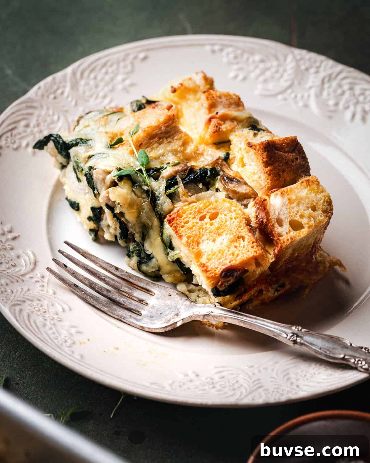 A slice of savory bread pudding with spinach, cheese, and mushrooms sits on a white decorative plate, accompanied by a fork. The dish features golden, toasted bread cubes and leafy greens.