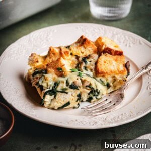A slice of savory bread pudding with spinach, cheese, and herbs sits on a decorative white plate next to a fork. The bread cubes are golden and toasted, and the dish is served on a rustic green table.