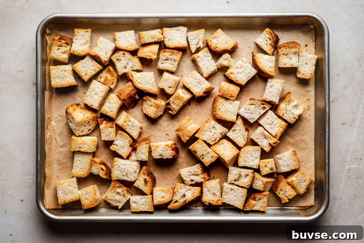 Cubed pieces of toasted bread are spread out on a parchment-lined baking sheet, ready to be used as croutons.