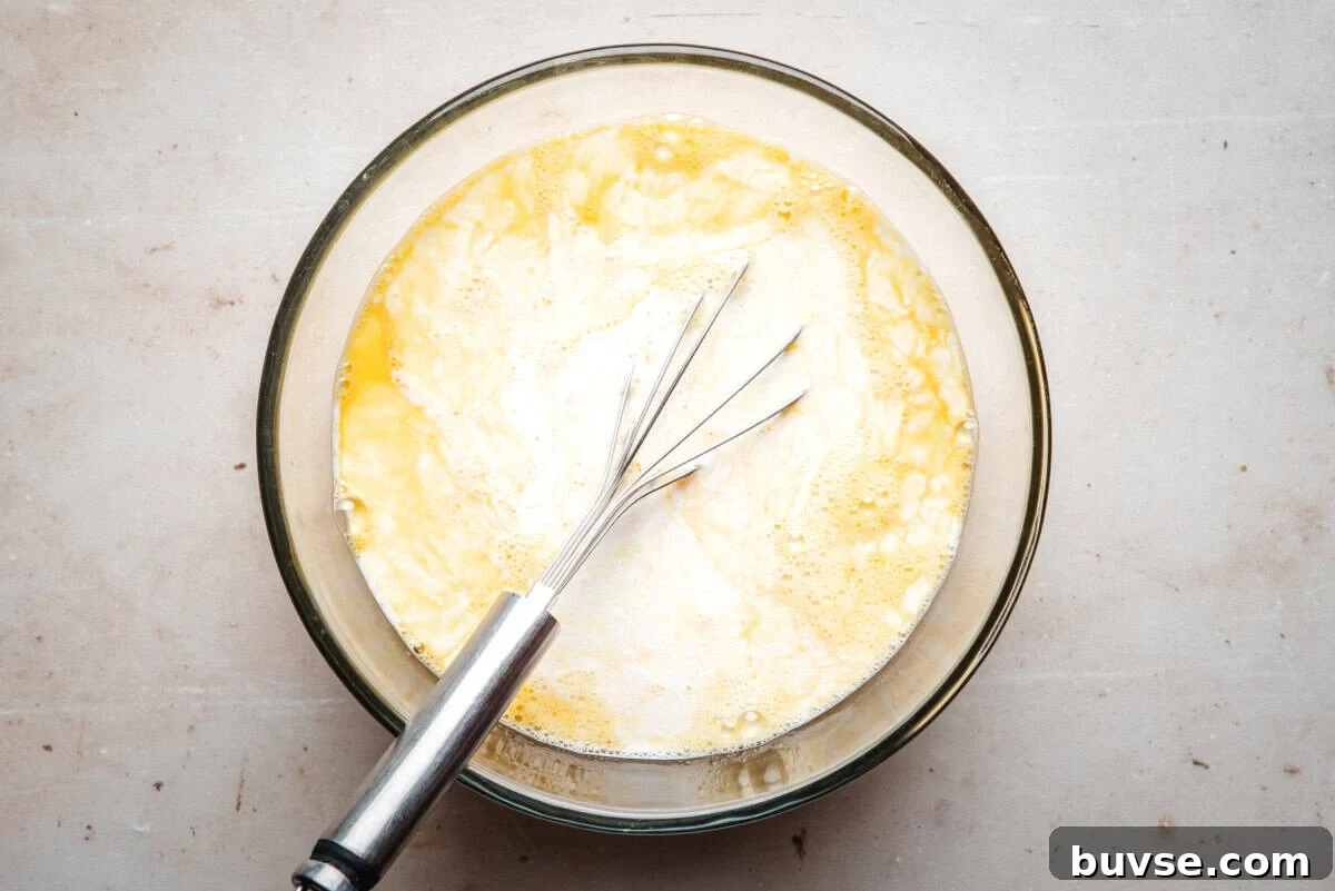 A whisk rests in a glass bowl filled with a mixture of eggs and milk, sitting on a light-colored countertop.