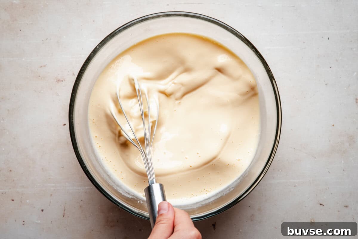 A hand holding a metal whisk is mixing a thick, creamy batter in a clear glass bowl on a light-colored surface.