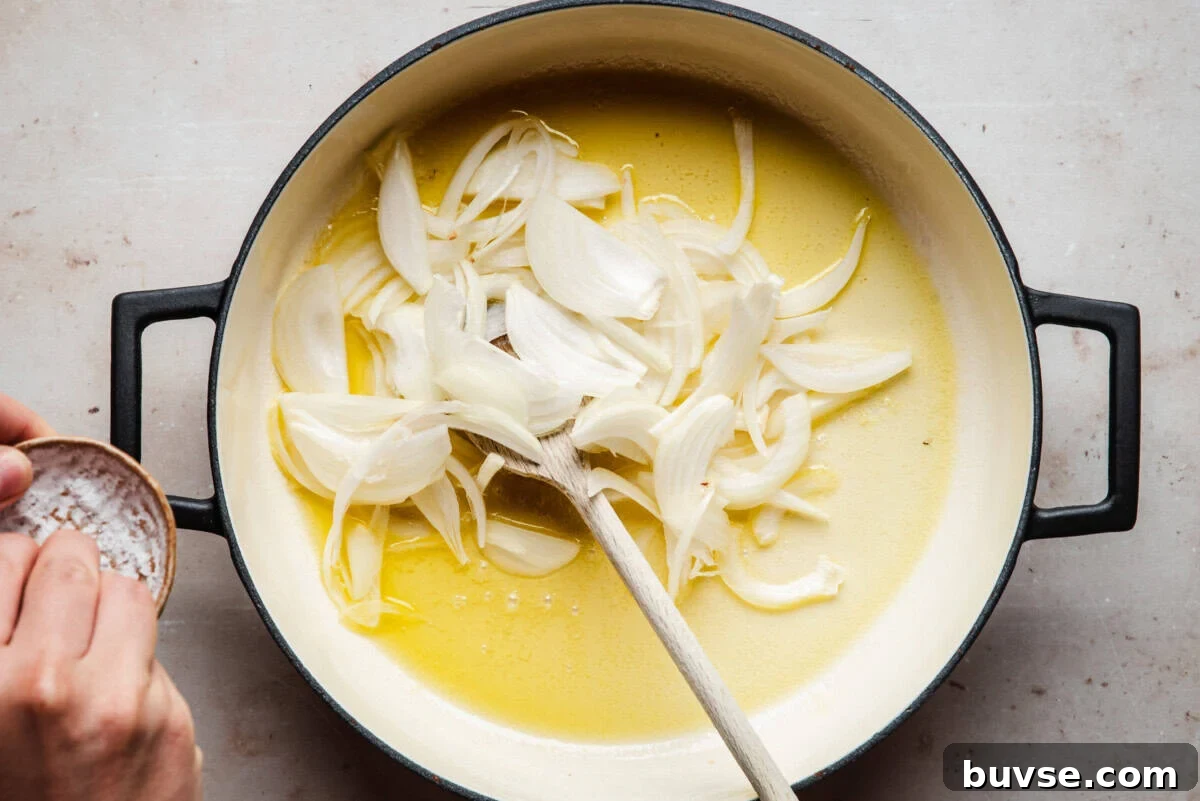 Sliced onions in a large pot with melted butter, being stirred with a wooden spoon. A hand holds a small bowl of salt over the pot, ready to season the onions.