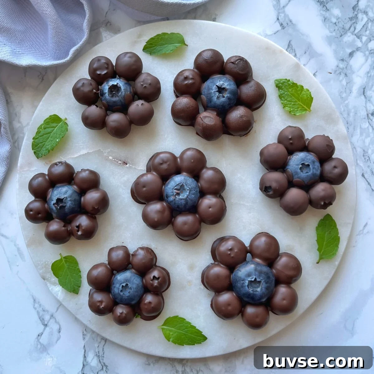 Homemade Chocolate-Covered Blueberries Recipe 2 Chocolate Covered Blueberry clusters arranged in a flower shape on a round marble plate, garnished with fresh mint leaves.