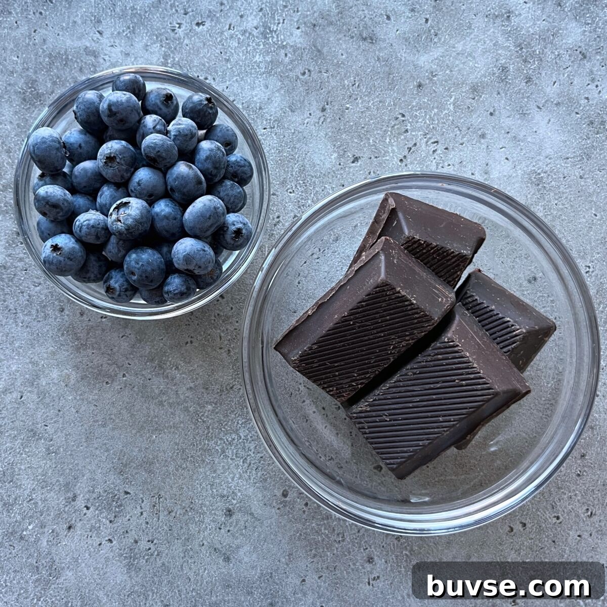 Homemade Chocolate-Covered Blueberries Recipe 4 Two glass bowls on a gray surface, one brimming with blueberries and the other holding four pieces of dark chocolate.