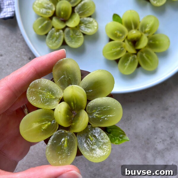 A hand holds a flower-shaped arrangement made from sliced green grapes, creating healthy bites displayed over a gray surface, with more grape flowers on a white plate in the background.
