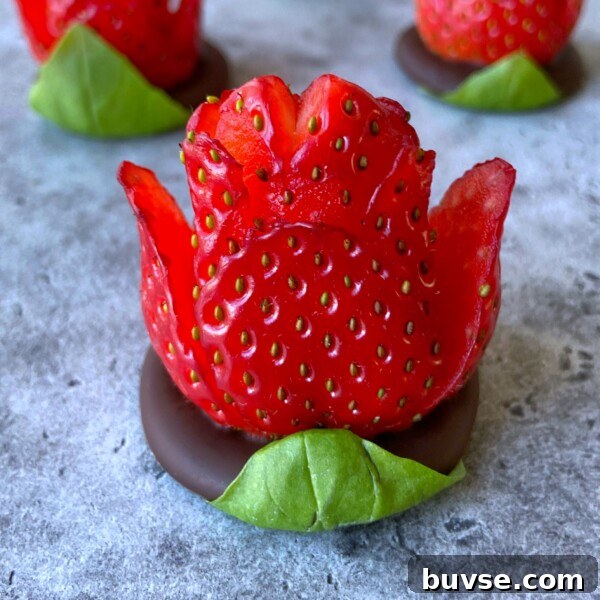 A chocolate strawberry rose, carved to resemble a blooming flower, sits atop a round piece of chocolate with a green leaf at the base, displayed on a gray surface. Other chocolate strawberry roses are softly blurred in the background.