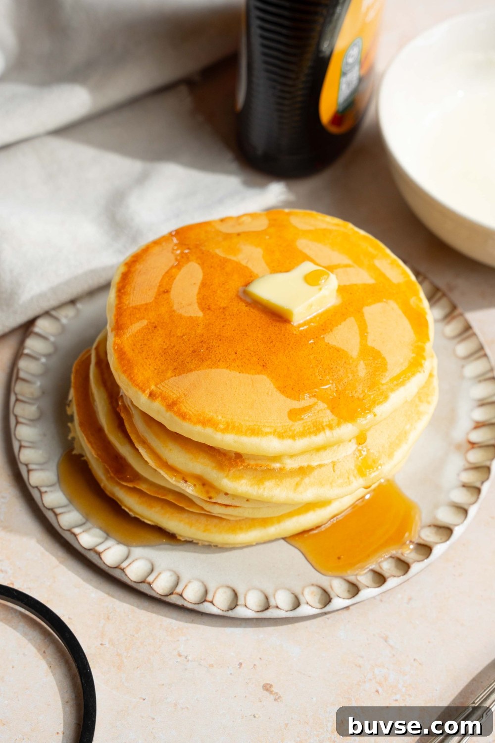 A white plate with a stack of pancakes with self rising flour topped with a pat of butter and drizzled with syrup. The plate is on a tan counter with a jar of syrup and a white cloth napkin.