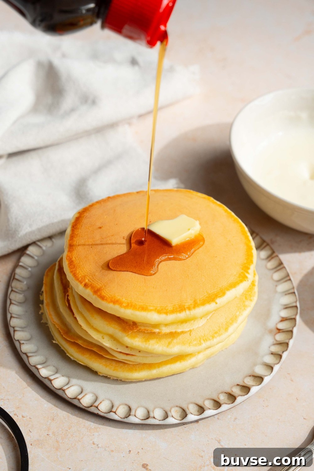 A white plate with a stack of pancakes with self rising flour topped with a pat of butter. Syrup is being poured over the stack. The plate is on a tan counter with a white cloth napkin.