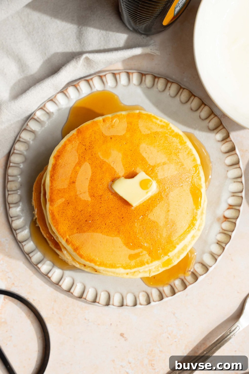 A white plate with a stack of pancakes with self rising flour topped with a pat of butter and drizzled with syrup. The plate is on a tan counter with a white cloth napkin.