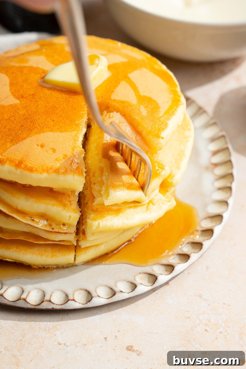 A white plate with a stack of pancakes with self rising flour topped with a pat of butter and drizzled with syrup. A fork is taking a bite from the stack. The plate is on a tan counter.