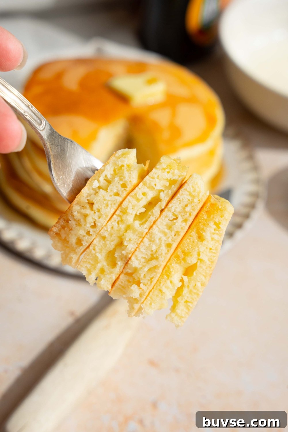 A close up of a fork with a bite of pancakes with self rising flour taken from the stack. There is a plate of pancakes on a tan counter blurred in the background.