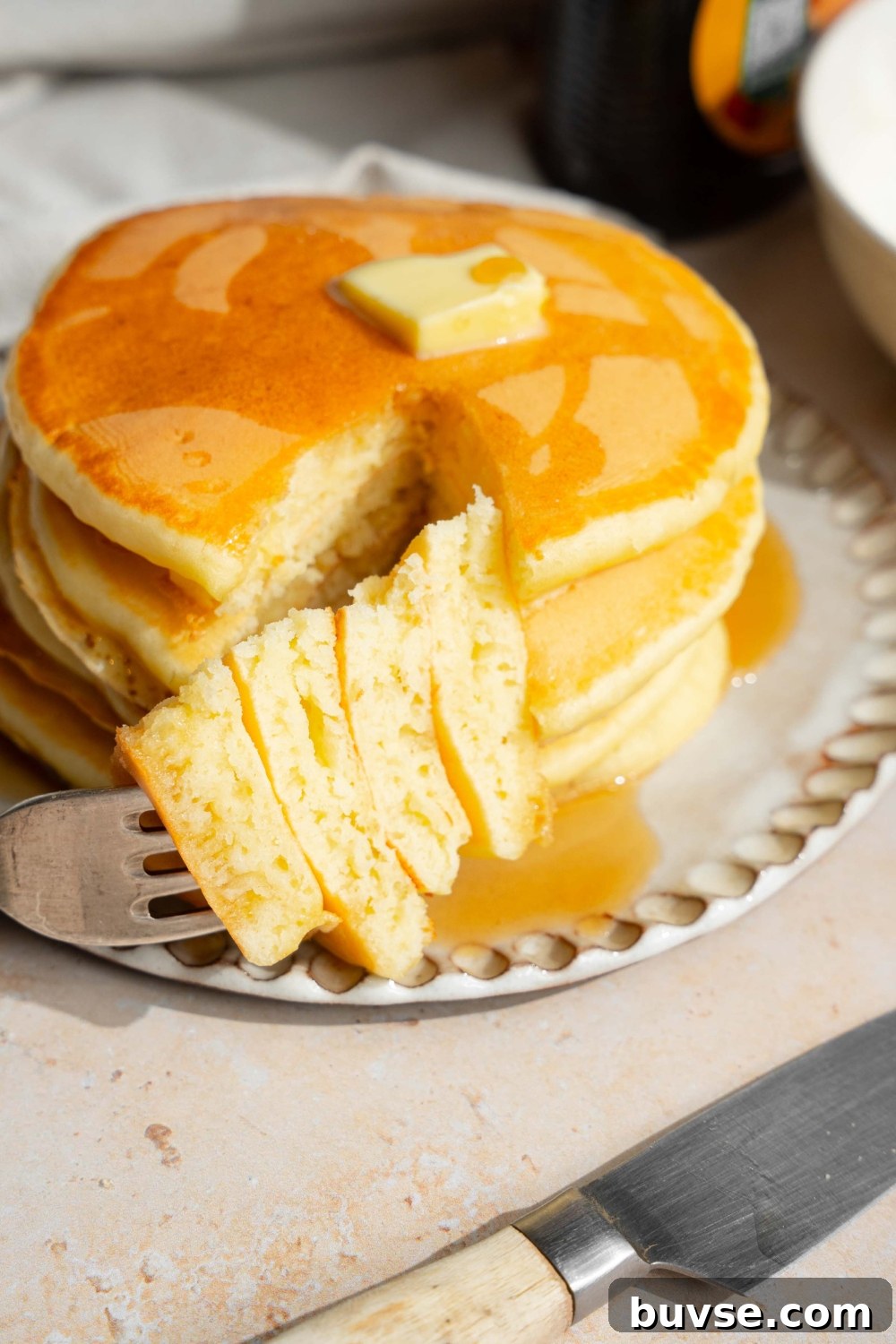 A white plate with a stack of pancakes with self rising flour topped with a pat of butter and drizzled with syrup. A fork is taking a bite from the stack. The plate is on a tan counter with a jar of syrup and a knife.