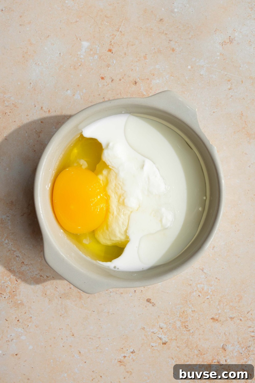 A white bowl with wet ingredients to make pancakes including egg and milk. The bowl is on a tan counter.