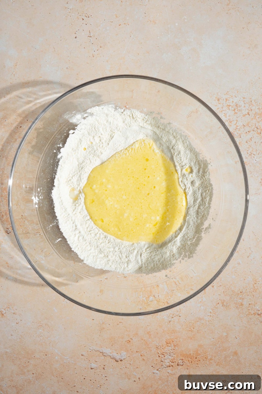 A large glass bowl with wet ingredients being added to dry ingredients for pancake batter. The bowl is on a tan counter.