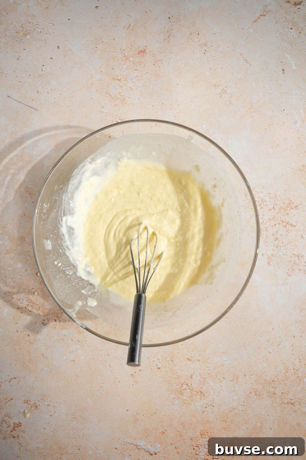 A large glass bowl with a whisk stirring pancake batter with self rising flour. The bowl is on a tan counter.