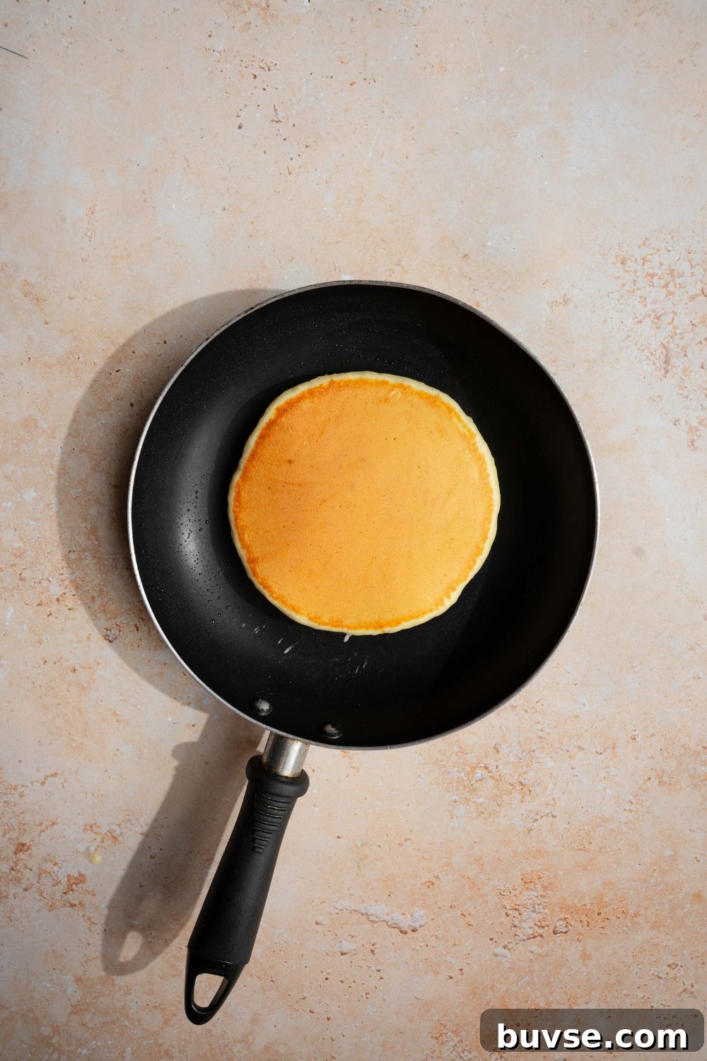 A skillet with a flipped pancake with self rising flour. The skillet is on a tan counter.