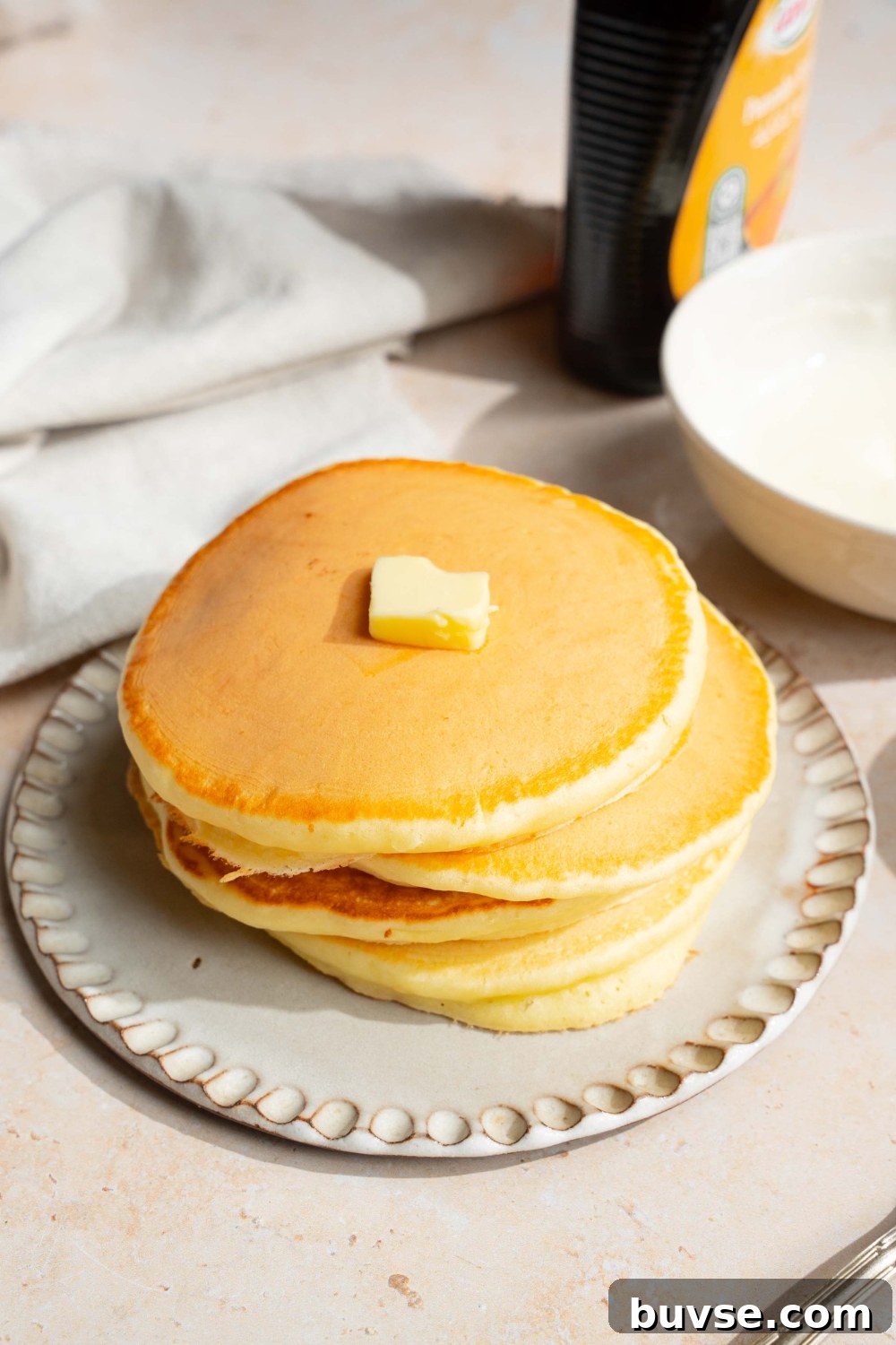 A white plate with a stack of pancakes with self rising flour topped with a pat of butter. The plate is on a tan counter with a jar of syrup and a white cloth napkin.