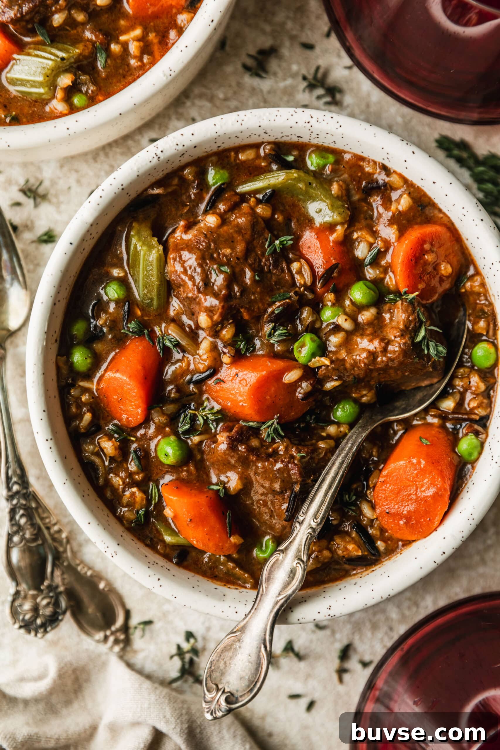Slow-Cooked Brisket Stew with Wild Rice and Vegetables 2 A white bowl of brisket beef stew with wild rice and vegetables on a beige counter next to red wine and a beige linen.