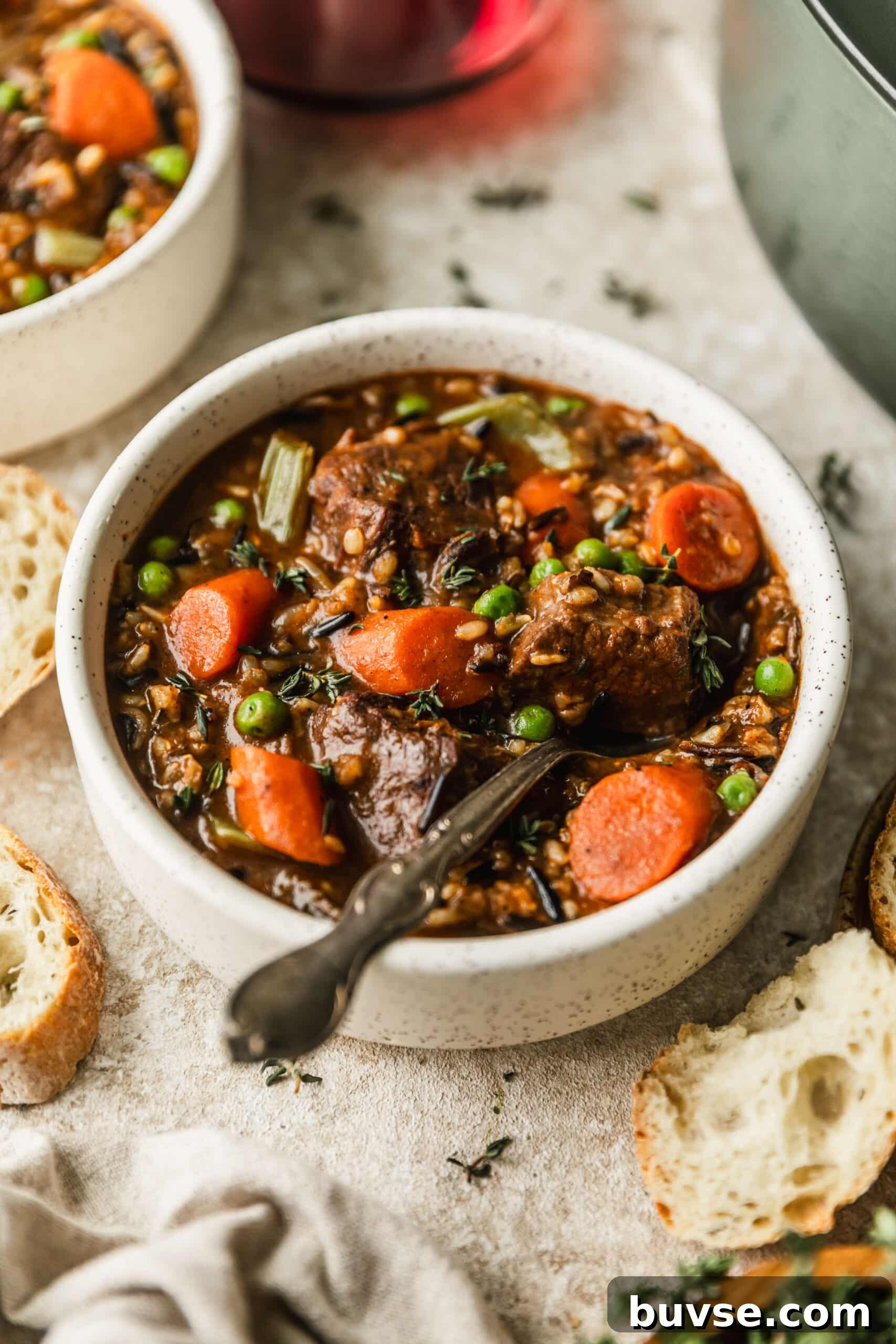 Slow-Cooked Brisket Stew with Wild Rice and Vegetables 8 Two white bowls of beef and wild rice soup on a tan counter next to bread, glasses of red wine, and a green Dutch oven with a beige background.