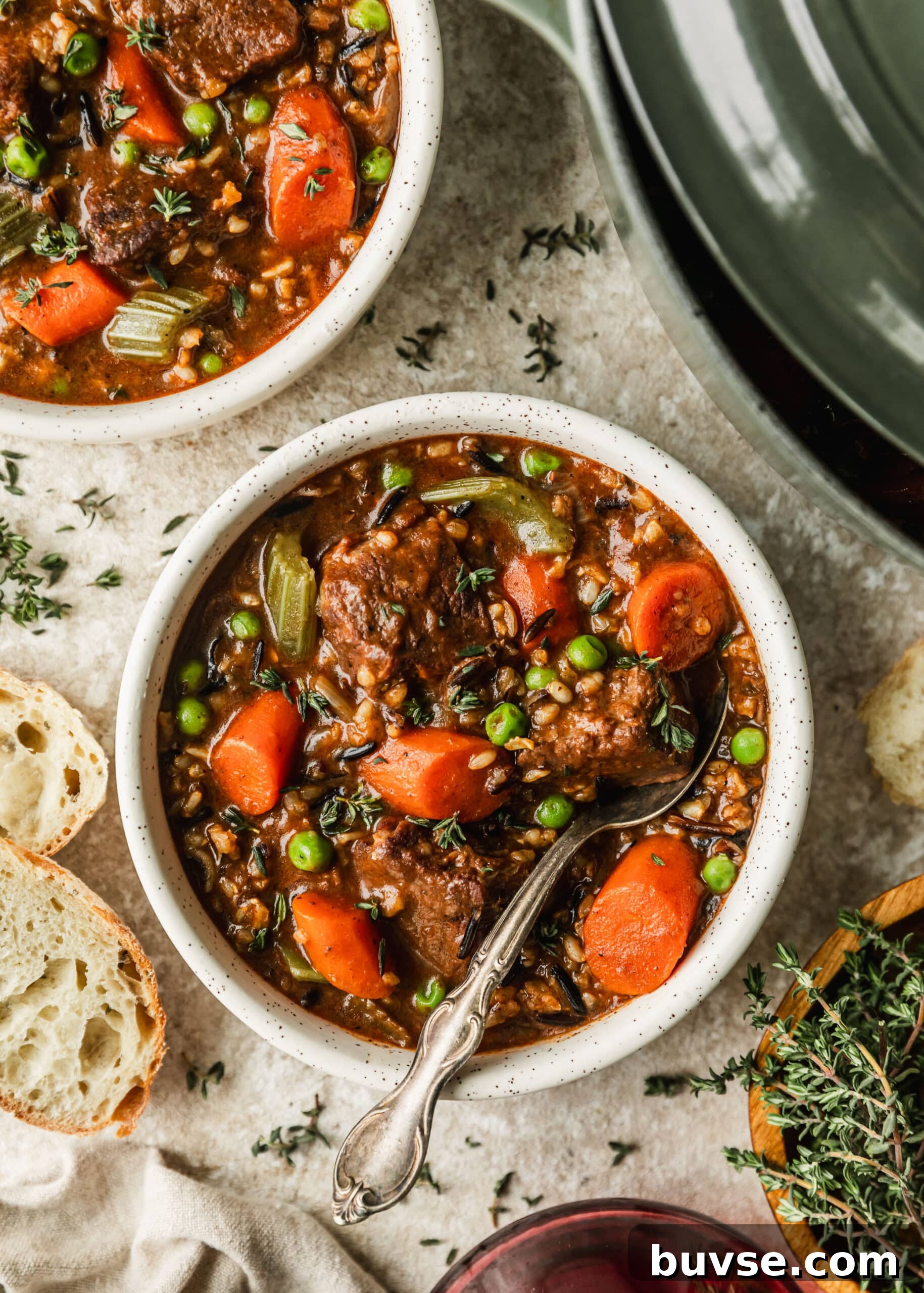 Slow-Cooked Brisket Stew with Wild Rice and Vegetables 9 Two white bowls and a green Dutch oven of brisket beef stew on a tan counter next to bread and red wine.