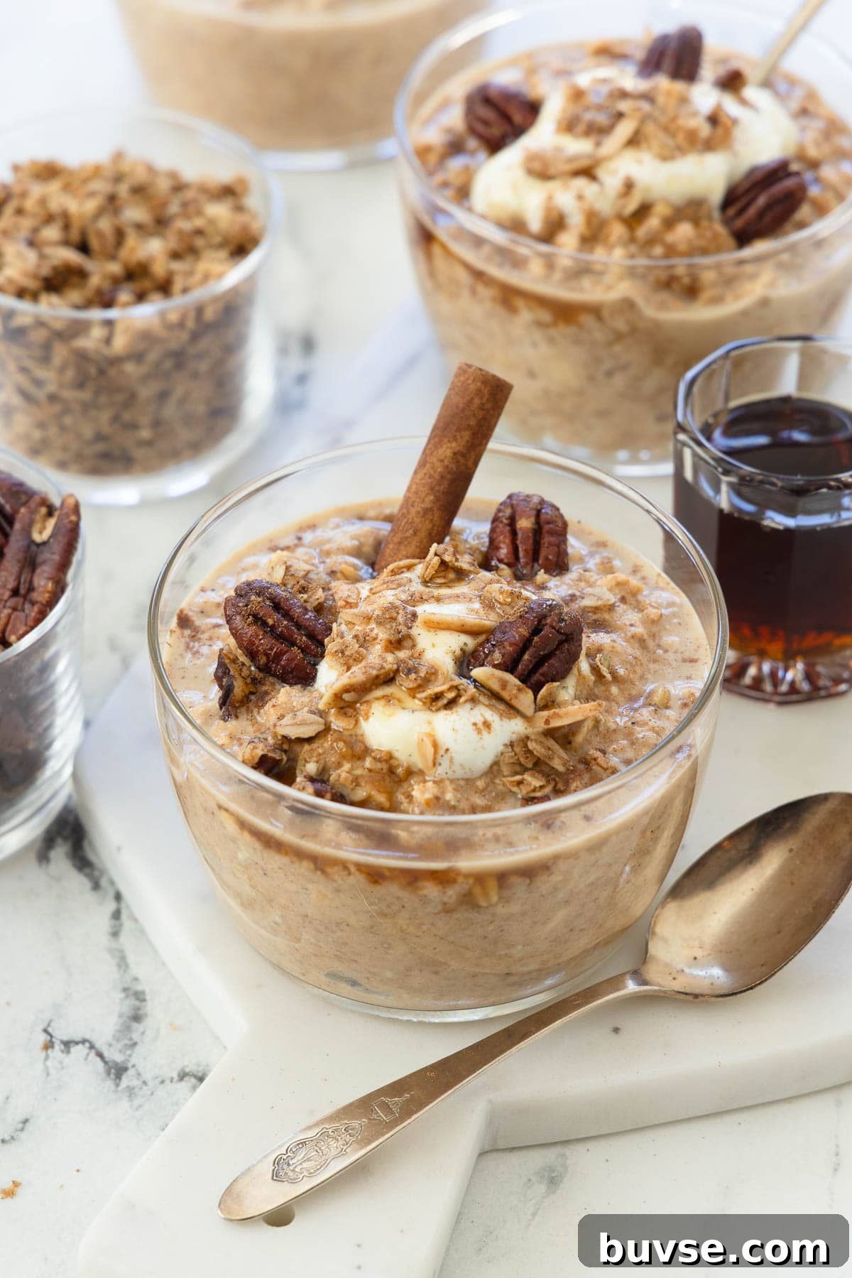 Bowls of pumpkin overnight oats with cinnamon stick, pecans, yogurt, and a silver spoon.