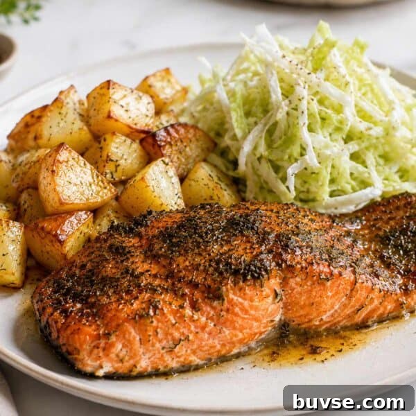 A photo of a sockeye salmon fillet cooked on a white plate, cooked with herbs on top. In the background of the white plate you have cubed potatoes and shredded cabbage salad