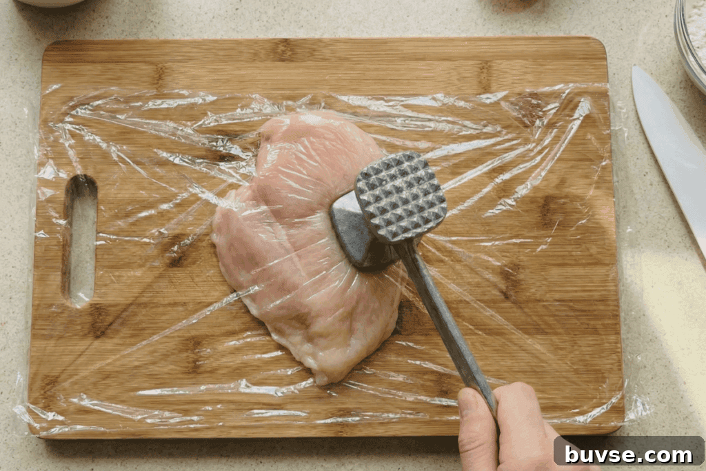pounding raw chicken on a wooden board with a meat mallet in between plastic sheets