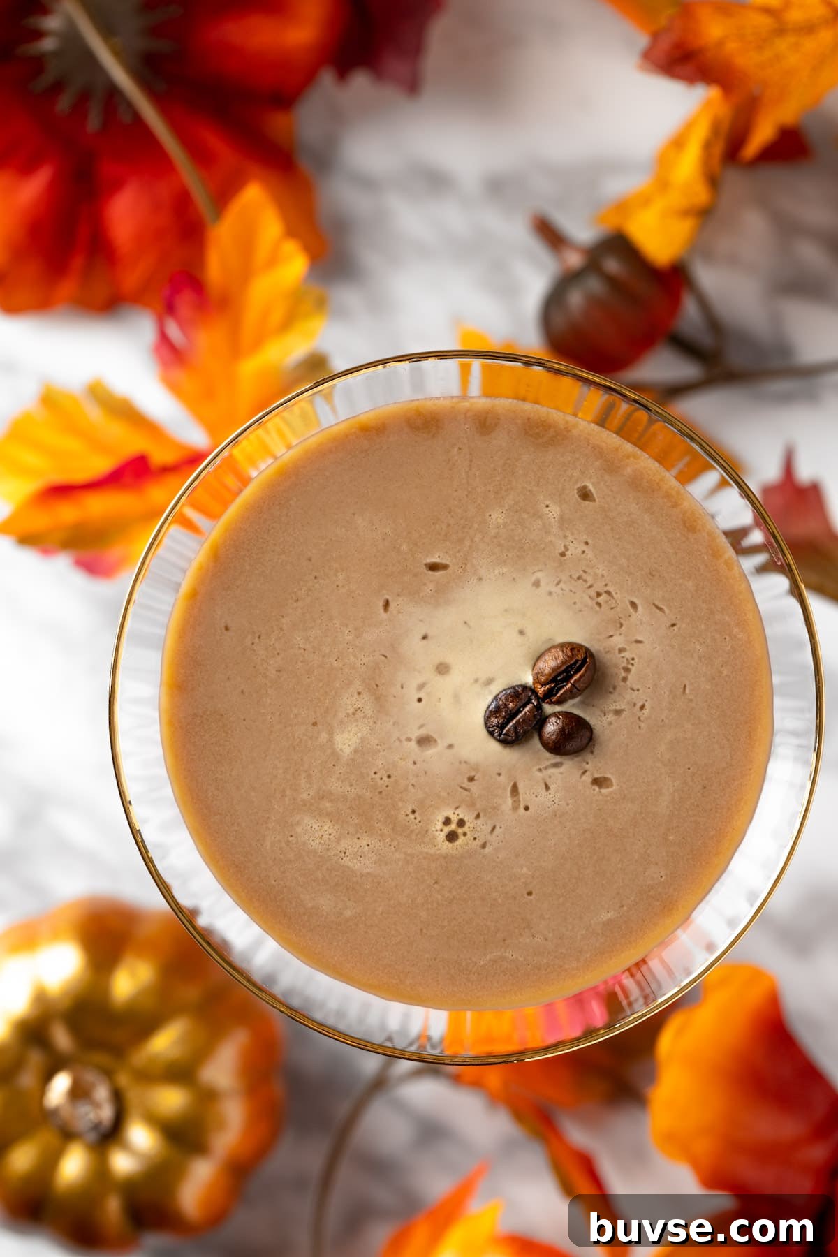 Pumpkin Spice Espresso Martini Cocktail Recipe 4 Overhead view of a pumpkin spice espresso martini on a white marble table next to pumpkins and leaves.