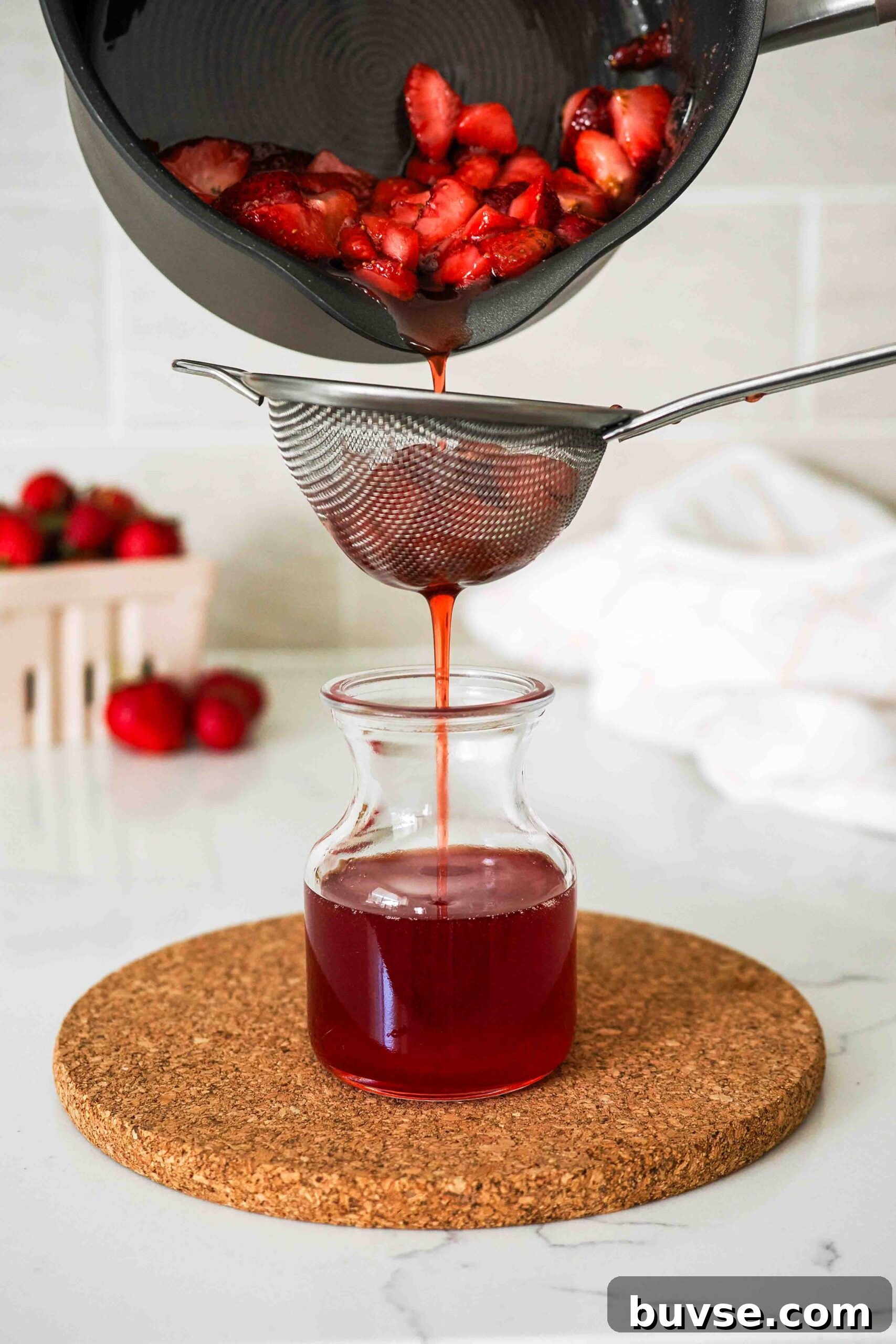 Strawberry syrup is poured out of a pot through a strainer and into a small jar.